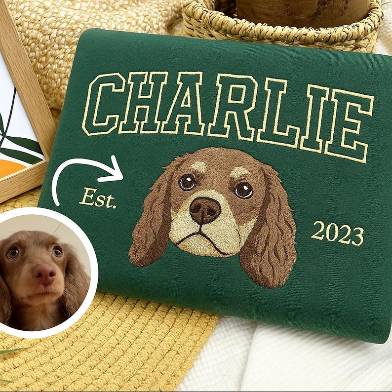 May include: A dark green sweatshirt with the name "CHARLIE" in gold lettering. Below the name is an embroidered dog face and the year "2023". A small photo of a dog is in the lower left corner. The word "Est." is next to an arrow.