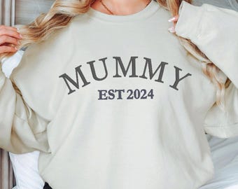 May include: A light beige sweatshirt with the text "MUMMY EST 2024" in black.