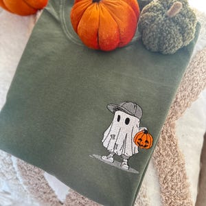 May include: An olive green sweatshirt with an embroidered cartoon ghost wearing a gray cap and holding an orange jack-o'-lantern. Two decorative pumpkins, one orange and one green, are placed on top of the folded sweatshirt. The image has a fall or Halloween theme.