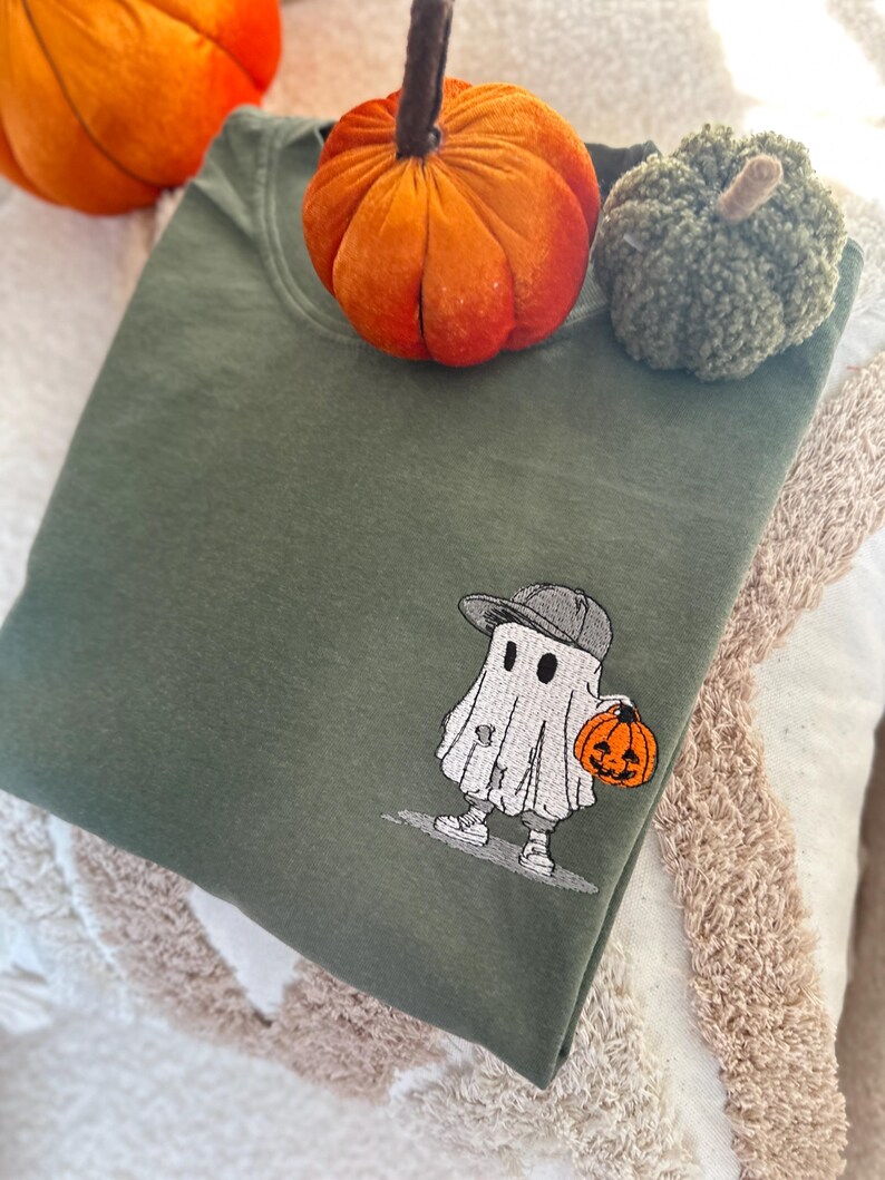 May include: An olive green sweatshirt with an embroidered cartoon ghost wearing a gray cap and holding an orange jack-o'-lantern. Two decorative pumpkins, one orange and one green, are placed on top of the folded sweatshirt. The image has a fall or Halloween theme.