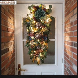 May include: A teardrop-shaped festive swag featuring green pine needles, eucalyptus leaves, and pinecones. It is decorated with metallic bronze and copper baubles, gold-toned snowflakes, and warm white string lights, creating a seasonal display.