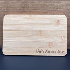 Könnte beinhalten: Ein rechteckiges Schneidebrett aus Holz mit einer natürlichen Holzmaserung. Das Brett hat den Text "Dein Wunschtext" in der rechten unteren Ecke eingraviert.