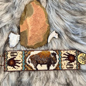 Può includere: Un braccialetto di perline con un designo di bisonte marrone e bianco, accenti turchesi e un'impronta di mano marrone su ogni lato. Il braccialetto ha una chiusura in ottone con un design a spirale.