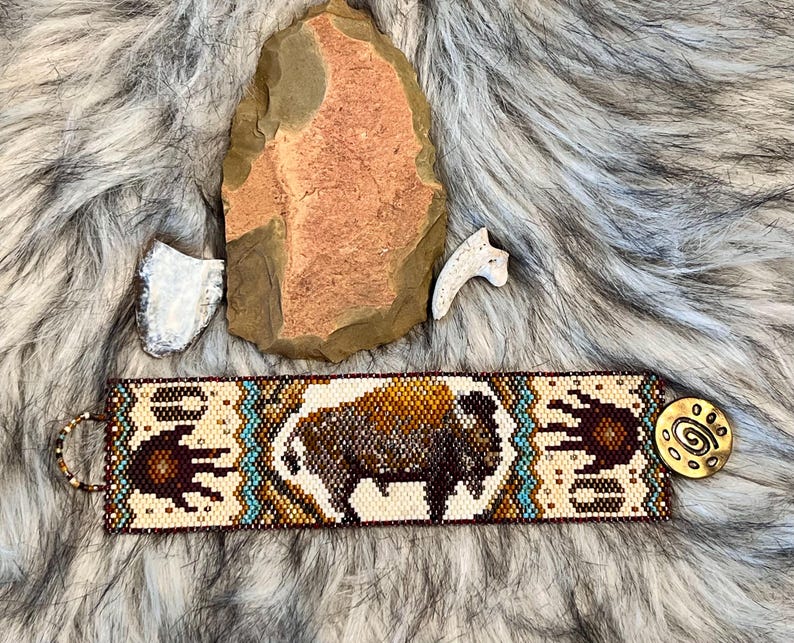 May include: A beaded bracelet with a brown and white bison design, turquoise accents, and a brown handprint on each side. The bracelet has a brass clasp with a spiral design.