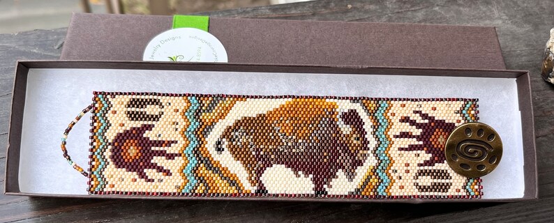 May include: A beaded bracelet with a brown and white bison design, turquoise accents, and a brown metal clasp with a swirl design. The bracelet is in a brown box with white tissue paper.