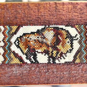 Op de afbeelding: Een rechthoekige kralenarmband met een centrale bizonafbeelding in bruin-, beige- en zwarttinten. Het ontwerp bevat handafdrukmotieven aan beide zijden, geaccentueerd met turquoise en crèmekleurige geometrische patronen, afgewerkt met een ronde knoop.
