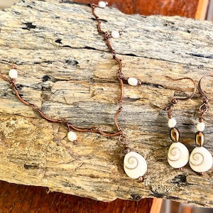 Puede incluir: Un collar y un par de pendientes a juego hechos de alambre de cobre. El collar presenta pequeñas perlas blancas y un colgante de concha en espiral. Los pendientes consisten en una pequeña perla, una cuenta metálica y una concha en espiral.
