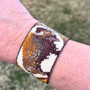 May include: A beaded bracelet with a brown, white, and gold design of a buffalo. The bracelet is made of small, colorful beads.