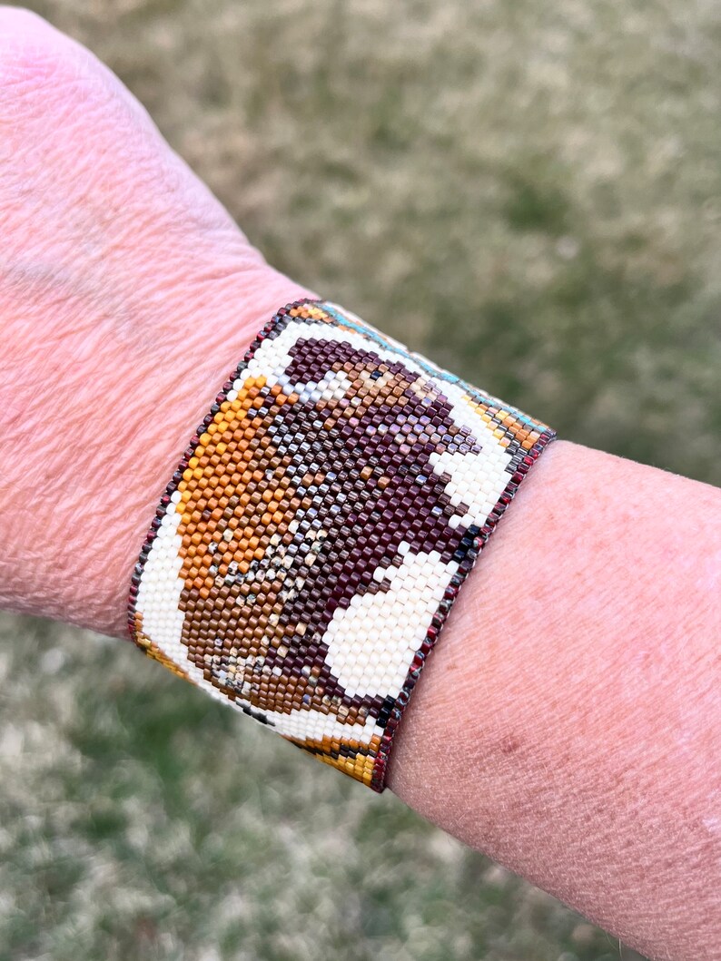 May include: A beaded bracelet with a brown, white, and gold design of a buffalo. The bracelet is made of small, colorful beads.