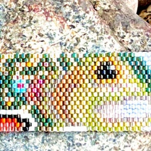 May include: A beaded bracelet with a colorful, intricate design. The bracelet features a pattern of small, multi-colored beads, including shades of green, yellow, orange, pink, and blue. The design appears to depict a stylized image. The bracelet is set against a textured background.