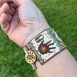 May include: A beaded bracelet with a brown bear paw print design, turquoise and gold accents, and a gold clasp with a spiral design.