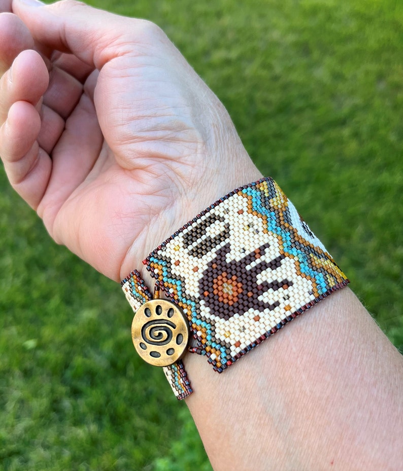 May include: A beaded bracelet with a brown bear paw print design, turquoise and gold accents, and a gold clasp with a spiral design.
