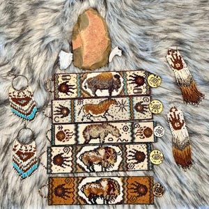 May include: Five beaded bracelets with Native American designs. Each bracelet features a different animal, including a buffalo, a horse, a bear, and two more buffalo. The bracelets are all beige with brown, blue, and white accents. The bracelets are displayed on a white faux fur background with two beaded earrings.