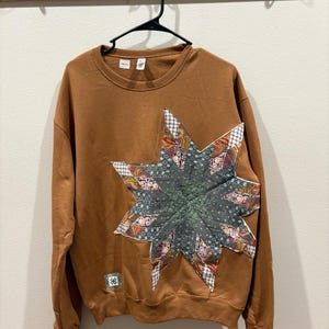 May include: A brown long-sleeve sweatshirt featuring a large, multi-pointed star appliqué on the front. The star is constructed from various fabric patches in floral and plaid patterns. A small square patch with a geometric design is near the hem.