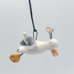 May include: A whimsical, handmade ceramic seagull ornament. The seagull is white with gray and yellow accents, and a small bird perched on its head. It is suspended by a black string, perfect for hanging.
