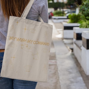 May include: Beige tote bag with the text "MY WISH MY COMMAND" in gold. The bag features gold line art of eyes, hands, and stars. The bag is carried over the shoulder of a person wearing a gray shirt and blue jeans.
