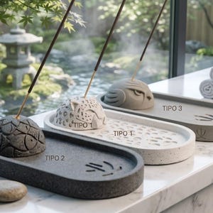 May include: Three oval incense holders with dome-shaped burners. The dark grey holder features a fish pattern, the white speckled holder is labelled TIPO 1, and the grey holder with an owl design is labelled TIPO 3. Each holds a thin incense stick.