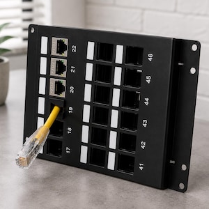 Può includere: Un pannello patch di rete in metallo nero con quarantotto porte numerate, dotato di spazi bianchi per etichette accanto a ciascuna porta. Un cavo Ethernet giallo con un connettore in plastica trasparente è inserito nella porta diciannove.