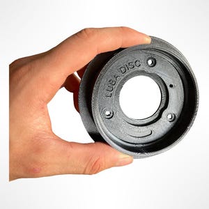 Peut inclure: Un objet circulaire gris avec le texte "LUBA DISC" imprimé sur le côté. L'objet comporte un trou central et plusieurs petits trous sur le bord. L'objet est tenu par une main sur fond blanc.