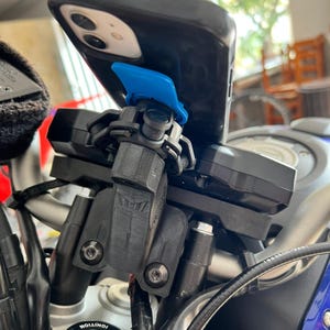 Op de afbeelding: Een zwarte smartphonehoes met een blauwe montageclip is bevestigd aan een zwarte motorstuurhouder. De houder is bevestigd met zilveren bouten. Het contactslot van de motor is zichtbaar.