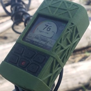 Puede incluir: Una unidad de control de detector de metales de mano con una funda protectora de color verde oliva texturizada con un patrón de rejilla geométrica. La pantalla digital muestra el número 76, el texto signal curve y varios ajustes.