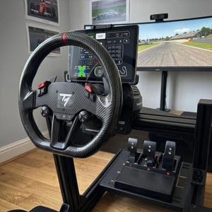 May include: A black carbon fiber racing steering wheel with a red top center stripe, mounted on a simulator rig. It features a central tablet displaying telemetry data, paddle shifters, and a set of three metal pedals positioned on the base below.