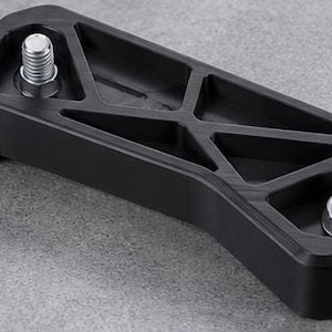 May include: A black, rectangular, rigid component featuring a geometric, truss-like cutout pattern on its top surface. Two silver-colored, threaded metal bolts with integrated nuts are positioned at opposite ends of the black frame.