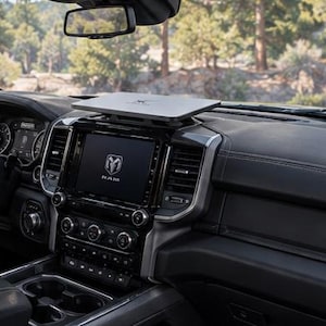Op de afbeelding: Het interieur van een Ram Laramie truck met een zwart dashboard, een centraal touchscreen met het Ram-logo, een stuurwiel met het Ram-embleem en een plat, rechthoekig grijs dienblad gemonteerd bovenop de dashboardconsole.