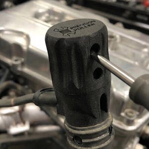 May include: A black, textured, cylindrical Precision Puller tool with a grooved exterior and multiple circular side openings. A metal screwdriver tip is inserted into one of the side holes of the tool, which is attached to a dark rubber hose.