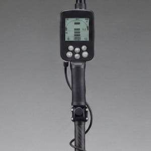 Puede incluir: Una caja de control de detector de metales negra montada en un eje vertical. La pantalla rectangular muestra una interfaz digital con cinco botones de control circulares debajo. Un cable negro recorre el lateral del eje hasta la unidad.