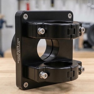 May include: A black metal mounting bracket featuring two circular clamps with silver hex bolts. The side of the rectangular plate is engraved with the text MONTURA PARA BASS SHAKER. The design includes a central circular opening and four corner bolts.