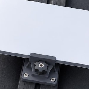 Pode incluir: Uma placa metálica retangular de cor prateada fixada a um suporte de montagem cinzento-escuro. O suporte possui um botão de ajuste central preto em forma de estrela e está fixado com quatro parafusos prateados num trilho preto ranhurado.