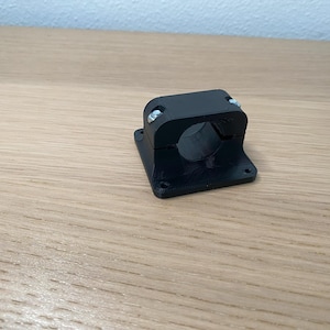 May include: Black plastic pipe clamp with a square base and two screws. The clamp is designed to hold a pipe or tube securely in place.