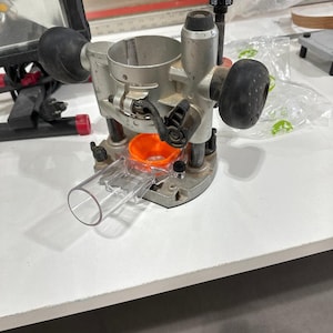 以下が含まれることがあります： 銀と黒のルーターテーブルで、透明なプラスチック製のダストコレクションチューブとオレンジ色のプラスチック製のインサートが付いています。テーブルには、白い文字盤に「0.8」と表示された黒いノブが付いています。