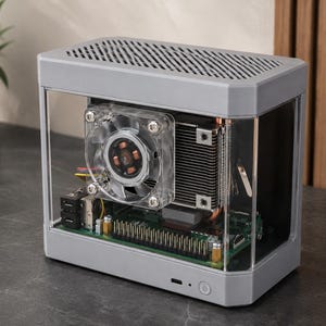 May include: A rectangular computer case with a light gray frame and clear acrylic side panels. The interior reveals a green circuit board, a metallic heat sink, and a transparent cooling fan. The top features a ventilated gray metal grille.