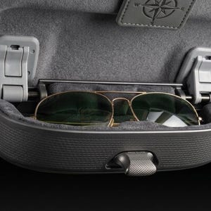 Puede incluir: Un soporte para gafas de techo en color gris oscuro con exterior texturizado e interior forrado de fieltro suave. El compartimento está abierto, mostrando unas gafas de sol tipo aviador con montura dorada. Se ve un pestillo con el texto PRESS TO OPEN.