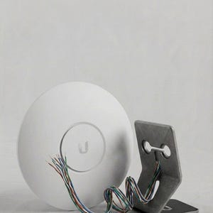 May include: A white circular wireless access point featuring a central U logo, positioned next to a gray metal mounting bracket. A bundle of multi-colored exposed electrical wires extends from the device, looping through the bracket's opening.