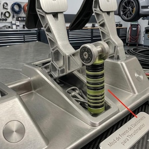 May include: A silver metal racing pedal set featuring a vertical stack of alternating black and lime green cylindrical elastomer dampers. A small rectangular plaque attached to the base displays the text Mod de Freno de Elastómero para Thrustmaster.