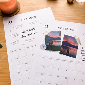 May include: Two open monthly calendar pages with handwritten notes and photos. One page is labeled "OKTOBER" and the other "NOVEMBER". A lit candle, stamps, and a glass of water are on a wooden surface.