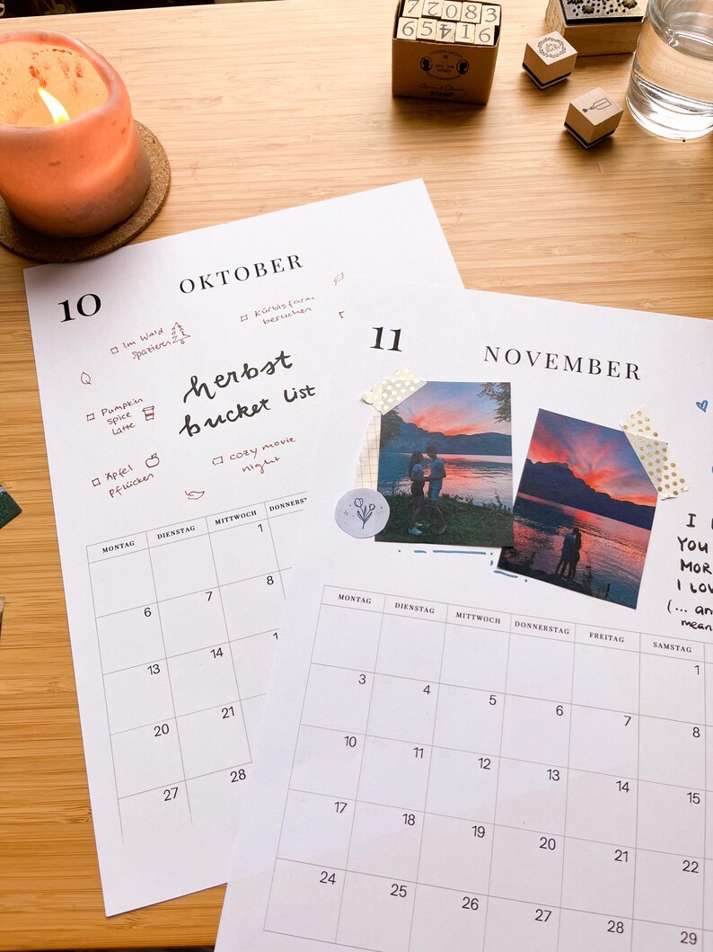 May include: Two open monthly calendar pages with handwritten notes and photos. One page is labeled "OKTOBER" and the other "NOVEMBER". A lit candle, stamps, and a glass of water are on a wooden surface.