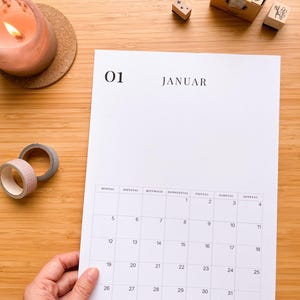 May include: A white calendar page with the month of January displayed, held by a hand. The calendar is on a wooden surface, with a lit candle, decorative tape, and number stamps nearby.