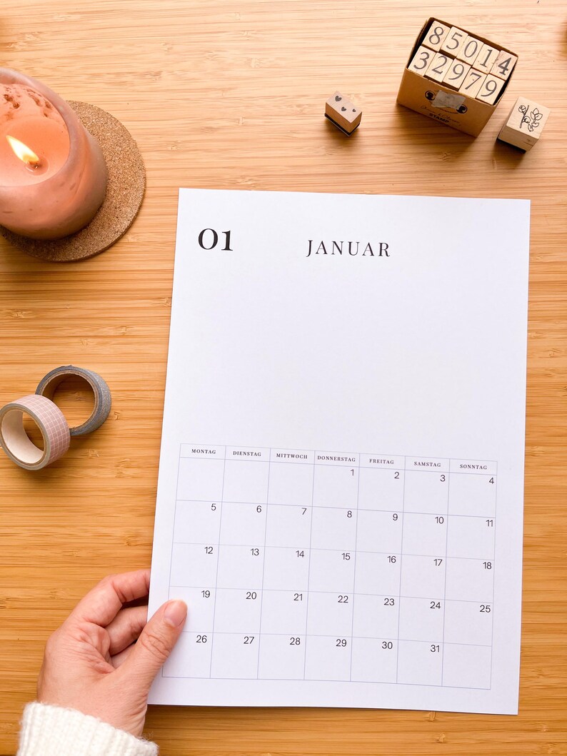 May include: A white calendar page with the month of January displayed, held by a hand. The calendar is on a wooden surface, with a lit candle, decorative tape, and number stamps nearby.
