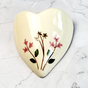 Puede incluir: Un plato de cerámica blanco en forma de corazón con un diseño floral. Las flores son rosas y amarillas con hojas verdes.
