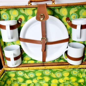 May include: A vintage picnic basket with a green and yellow fruit-patterned lining. The basket contains white plates and mugs secured with brown leather straps. The basket is made of woven wicker.
