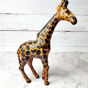 May include: A handcrafted giraffe figurine with a long neck and spotted pattern. The giraffe is primarily tan with black spots, and the ears are a lighter shade of tan. The figurine is standing on a white and gray surface.