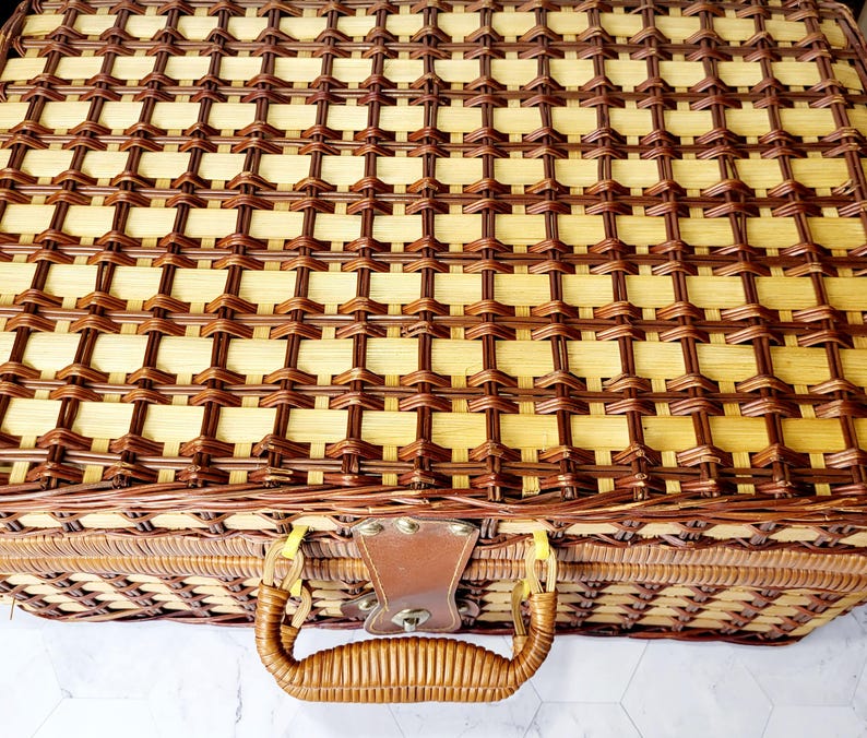 May include: A woven picnic basket with a brown leather handle and clasp. The basket is made of light and dark brown woven material. The handle is curved and attached to the basket with metal hardware.