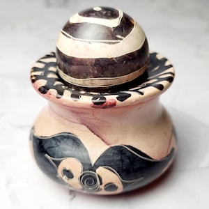 May include: A decorative ceramic jar with a lid. The jar is light pink with dark grey and black abstract designs. The lid is a dark brown sphere with white stripes. The jar is likely used for storage or decoration.