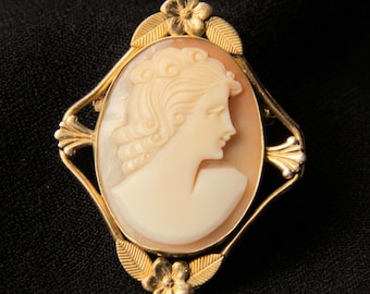 Peut inclure: Une broche en camée dorée avec un camée blanc représentant le profil d'une femme. Le camée est serti dans un cadre doré avec des accents floraux.