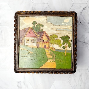 May include: A wooden box with a carved design around the edges. The lid features a painted scene of a house with a red roof and a green lawn.