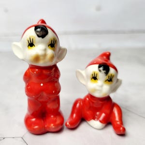 Puede incluir: Dos figuritas de elfo de cerámica roja y blanca. Un elfo está de pie con los brazos cruzados y el otro está sentado con las piernas cruzadas. Ambos elfos tienen el pelo negro, los ojos amarillos y la cara blanca.